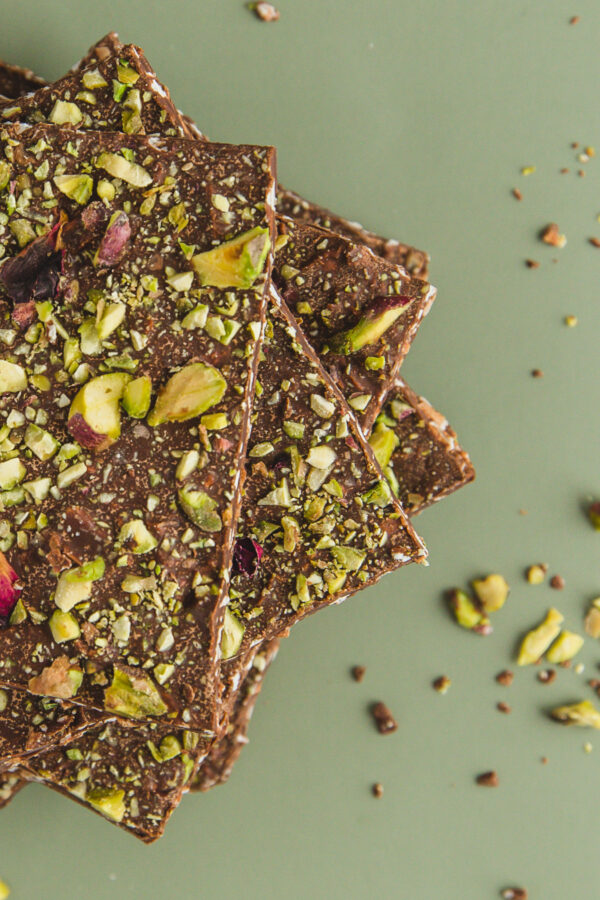 Vegan organic chocolate pistachio rose bark stacked in square pieces on a light green background with pistachios strewn beside it